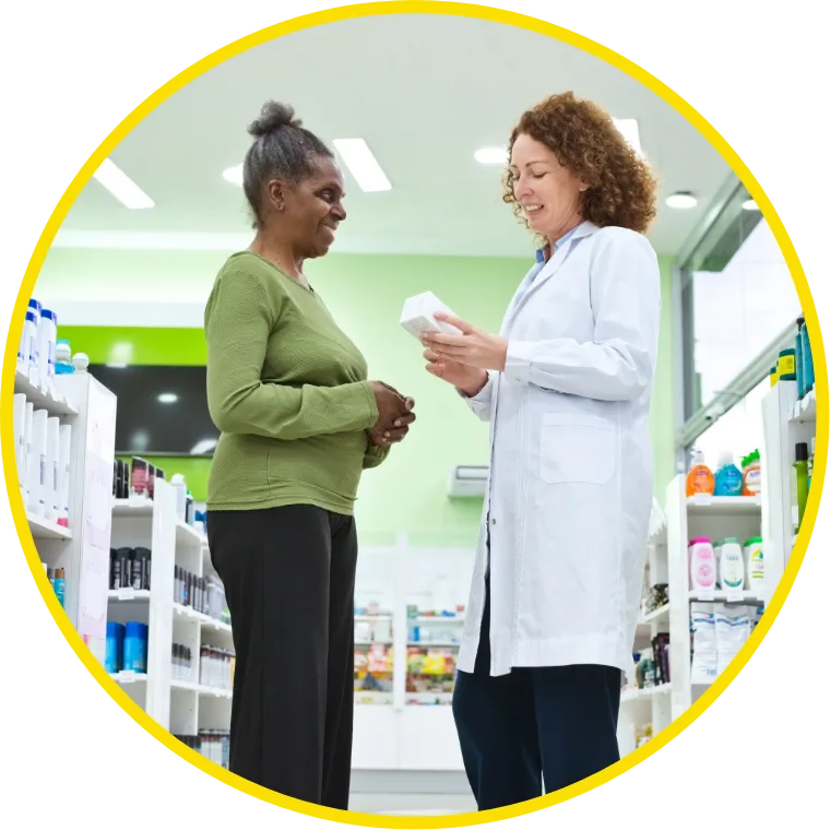 A pharmacist in a white coat discusses medication with a customer in a green sweater inside a bright pharmacy. Shelves of products surround them, conveying a helpful atmosphere.